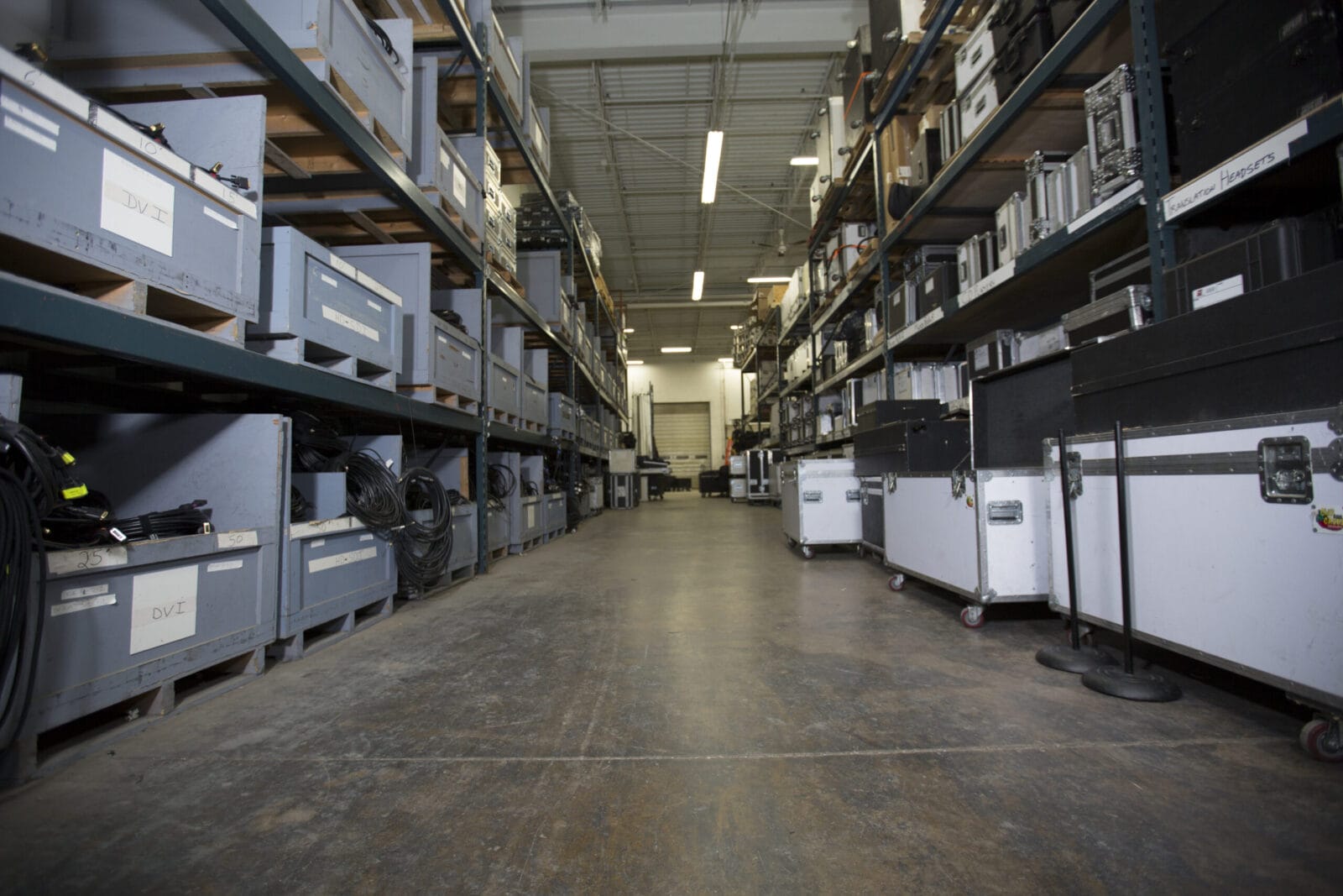 Warehouse aisle with shelves of equipment cases and storage bins.