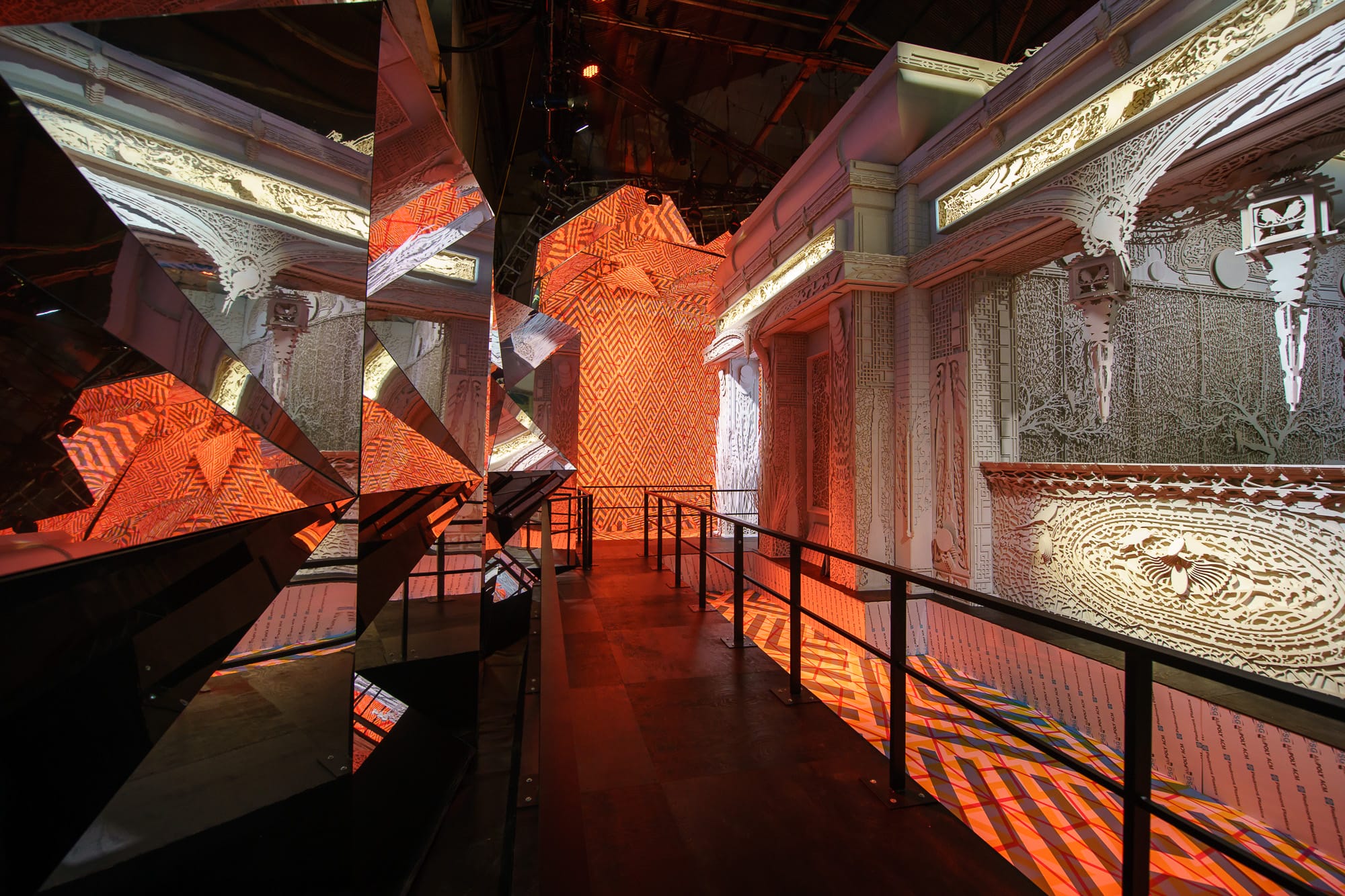 Hallway with mirrored walls reflecting ornate, patterned architecture and orange light.