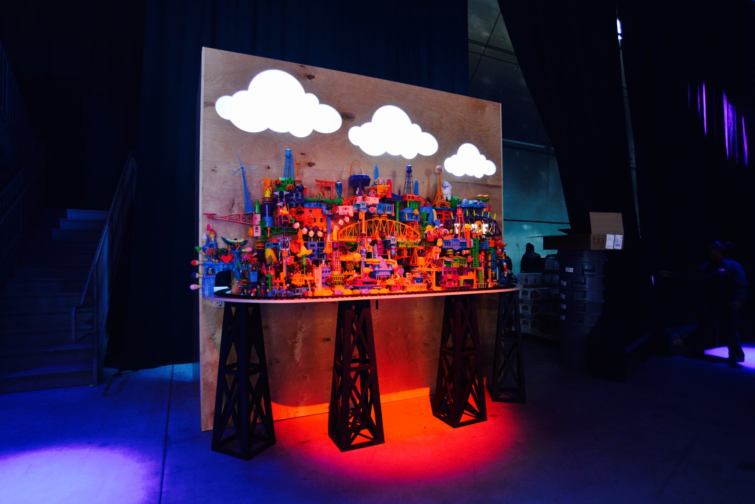 Colorful Lego cityscape with white clouds on a wooden backdrop, lit by red and blue lights.
