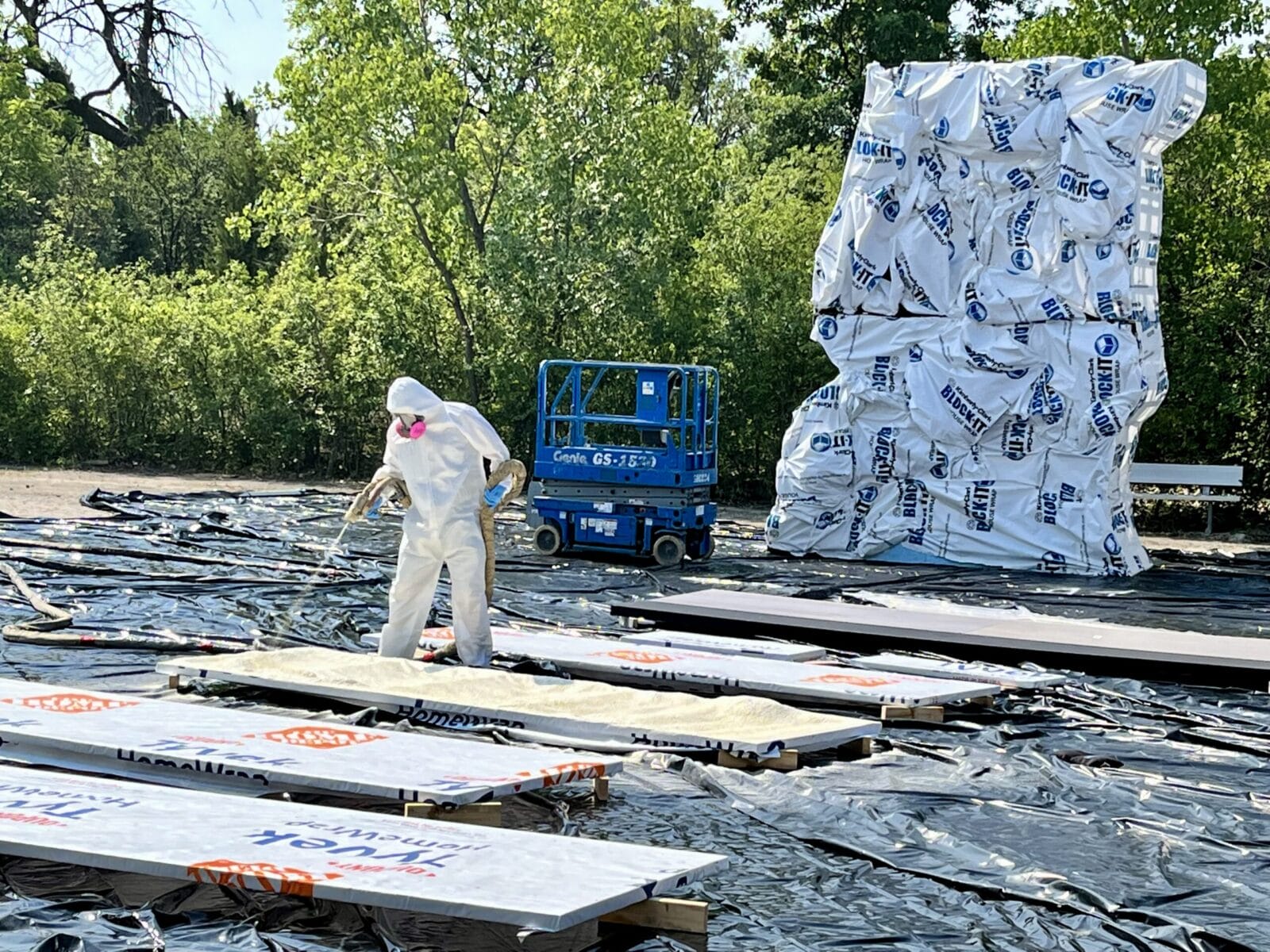 Worker in hazmat suit sprays insulation onto panels next to large wrapped structure.