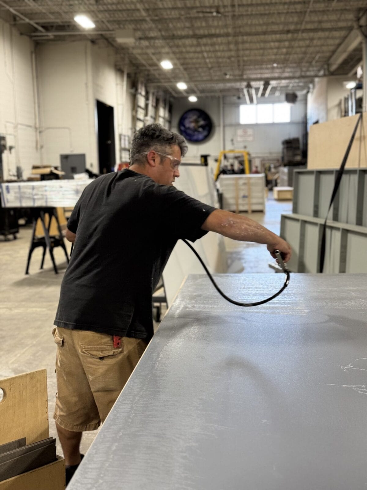 Man in safety glasses spray painting a large gray surface in a workshop.