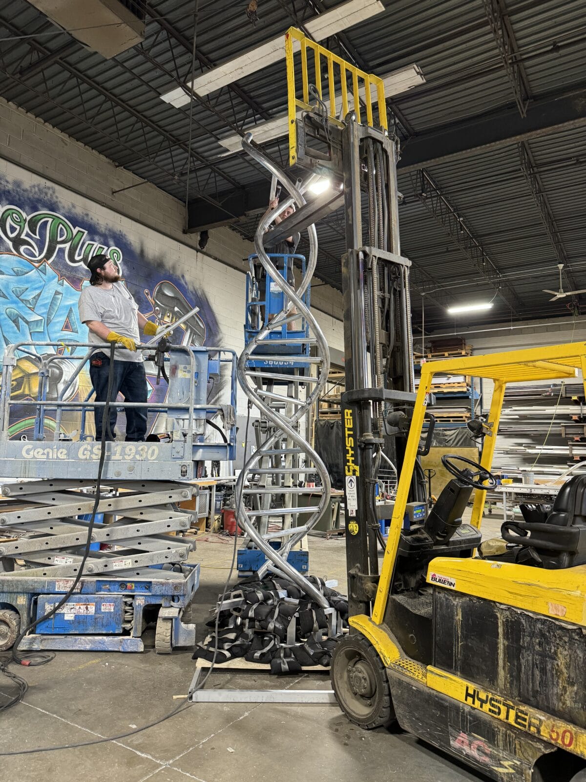 Workers install a large DNA sculpture using a scissor lift and forklift.
