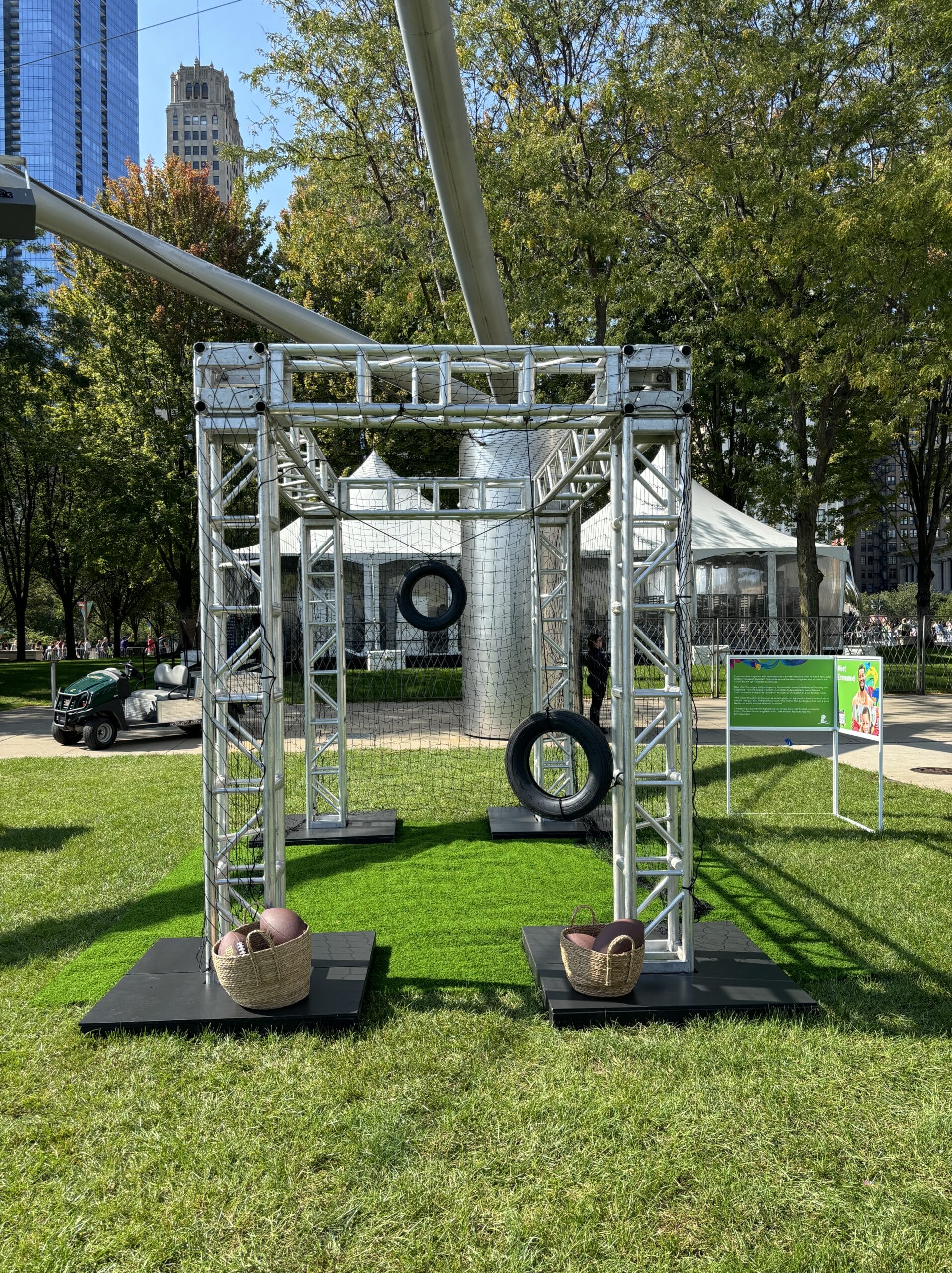 Metal truss structure with tires and baskets on artificial turf in a park.