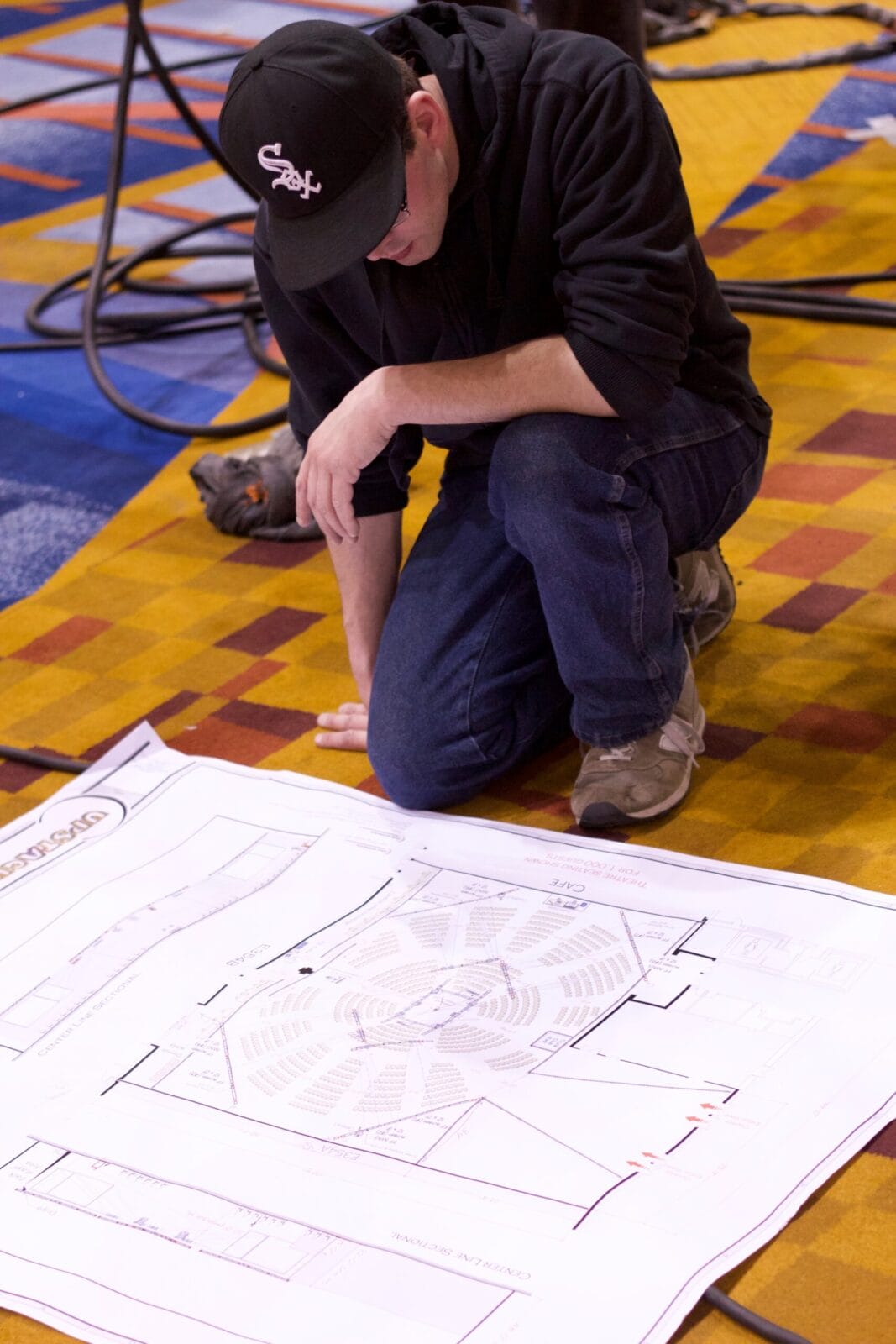 Man in black cap and hoodie studies blueprints on floor.