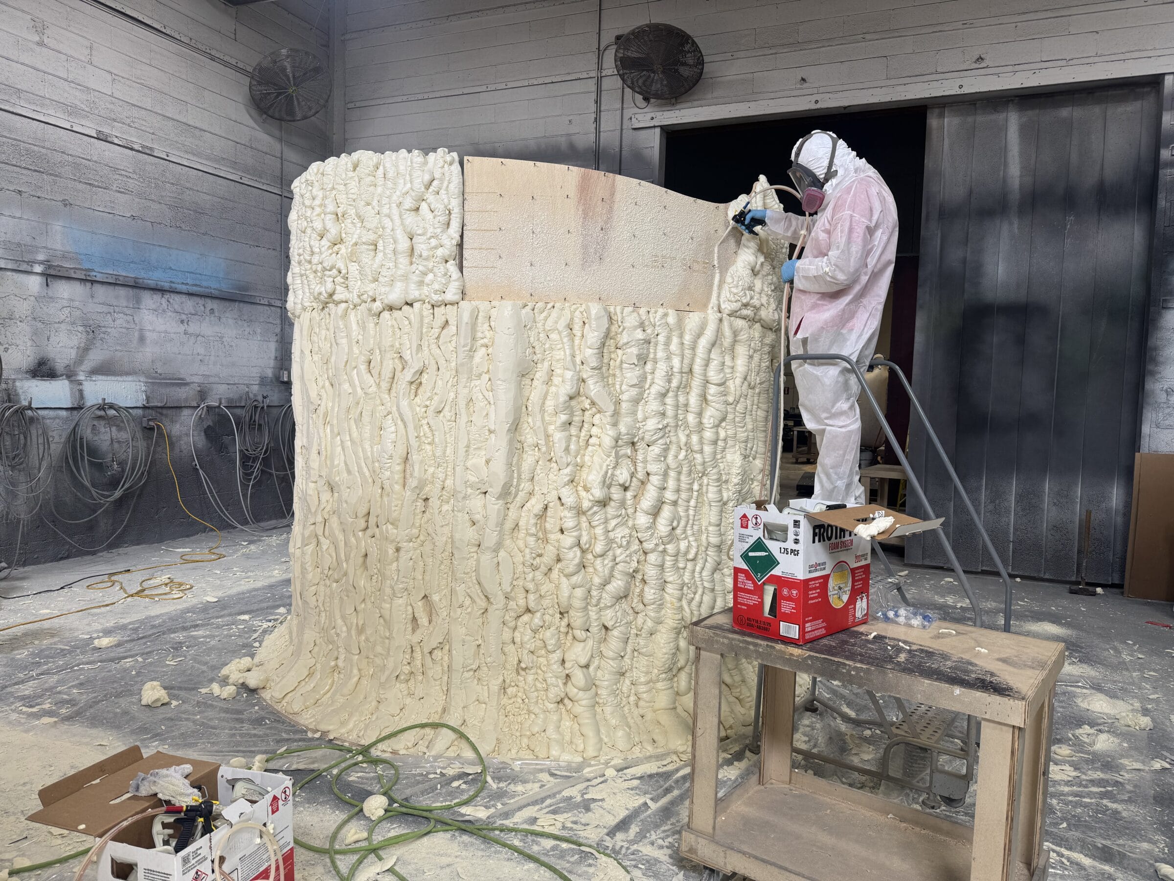 Worker in protective gear sprays foam onto a large, textured cylindrical structure.