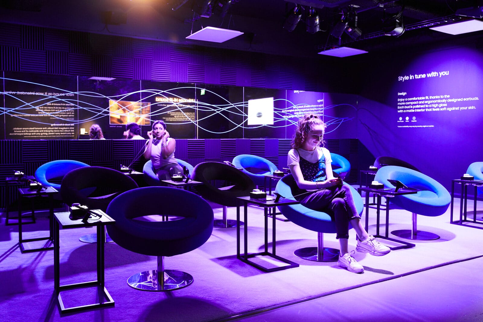 Two women sit in blue chairs in a room with purple lighting, wearing headphones.