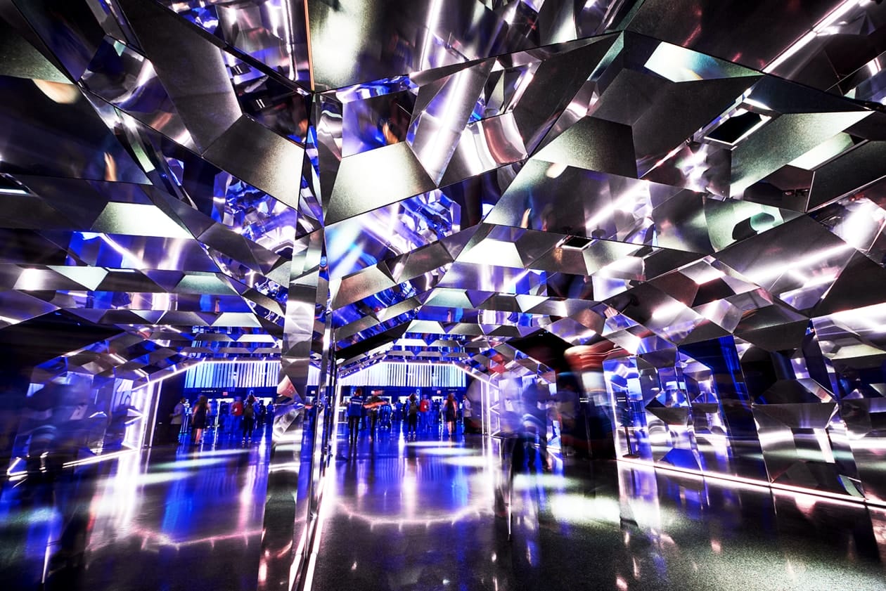 Hallway with mirrored, faceted walls reflecting blue light and people.