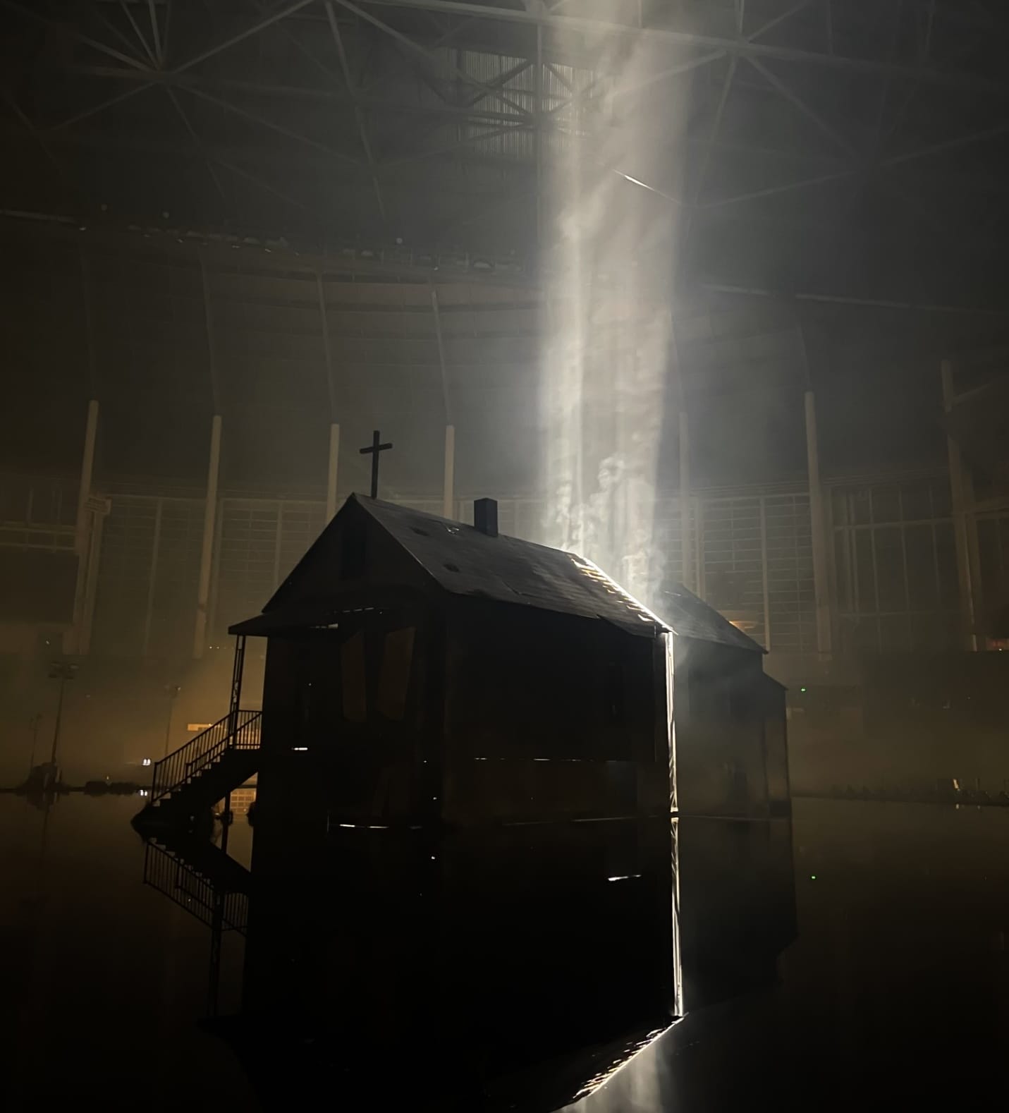 Dark, rustic cabin with a cross on roof, illuminated by dramatic light beams.