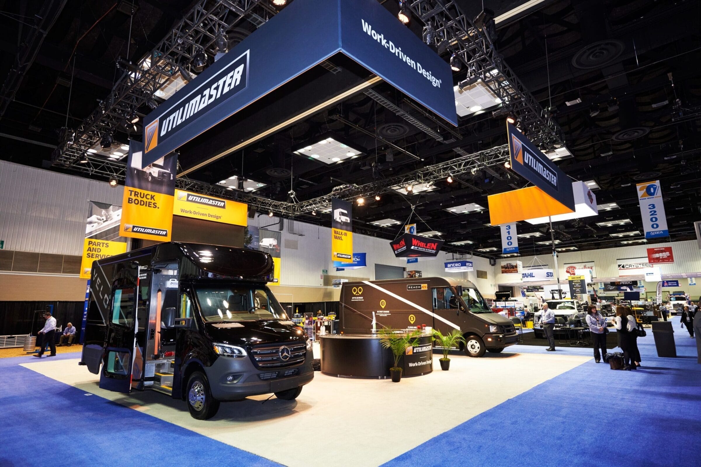 Black Utilimaster vans on display at a trade show with hanging banners.