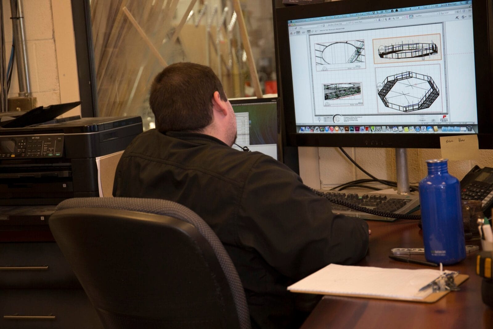 Man in black shirt working on computer with architectural drawings.