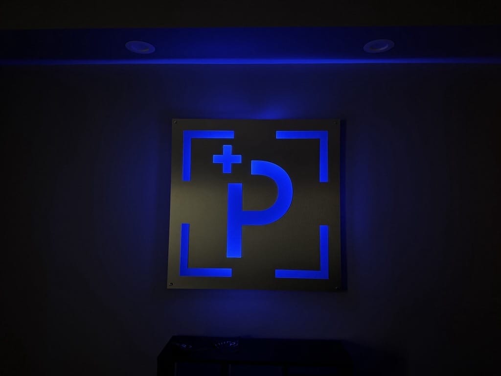Illuminated blue 'P' logo with a plus sign inside a square frame on a dark wall.